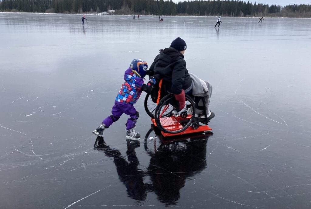 Un homme sur un fauteuil roulant et sur un Lugicap Ice Perf est poussé par un enfant qui patine sur un lac gelé / A man on a wheelchair which is on a Lugicap Ice Perf is pushed by a child ice-skating on a frozen lake