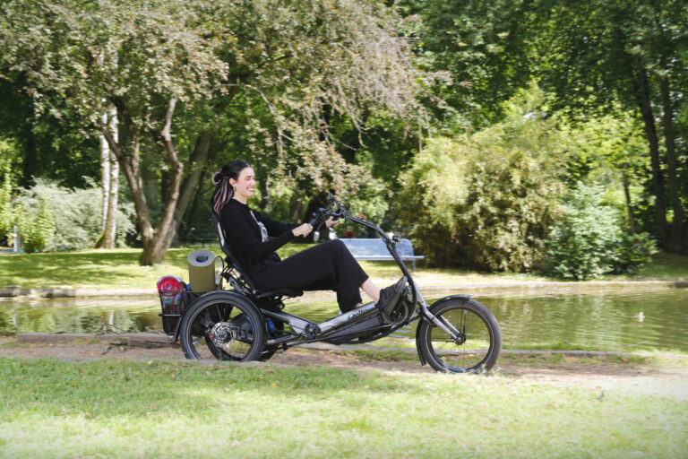 Français: Une femme est sur un vélo à trois roues. Elle est sur une chemin en poussière de roche dans un parc et devant un bassin d'eau. Elle avance en direction de la droite de l'image et a un grand sourire. English: A woman is on a three-wheeled bicycle. She is on a rock dust path in a park and in front of a water pond. She is moving towards the right of the picture and has a big smile on her face.