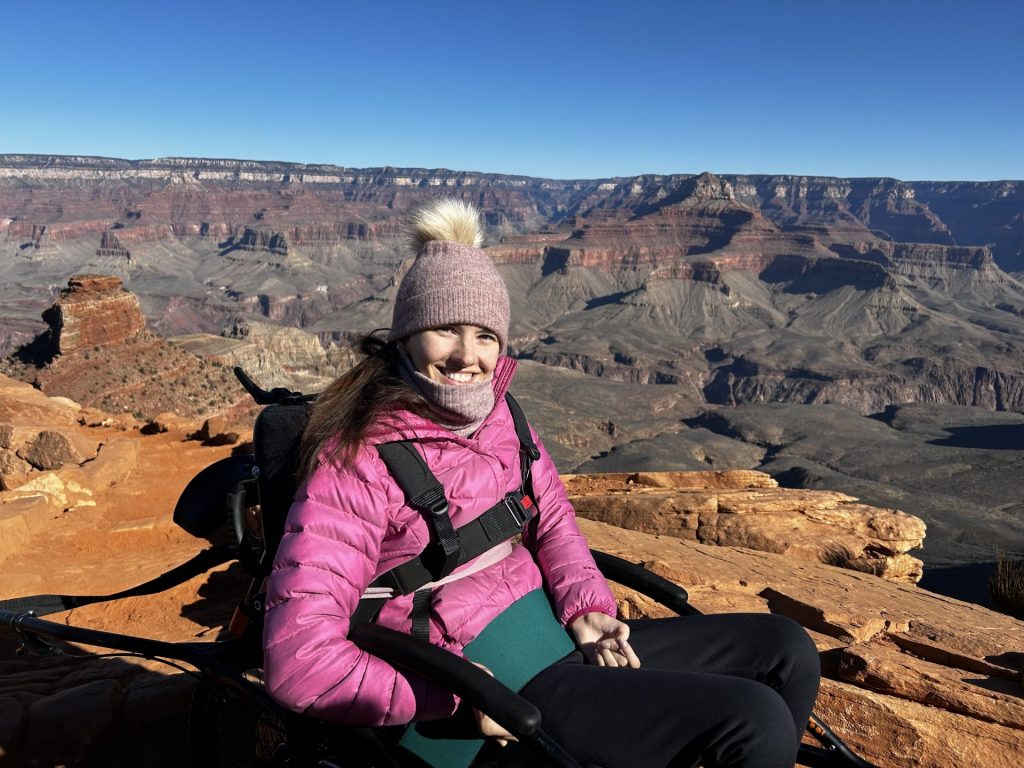 Français: Une jeune femme souriante est assise sur une Joëlette devant le paysage du Grand Canyon par beau temps. English: A smiling young woman sits on a Joëlette in front of the Grand Canyon landscape on a sunny day.