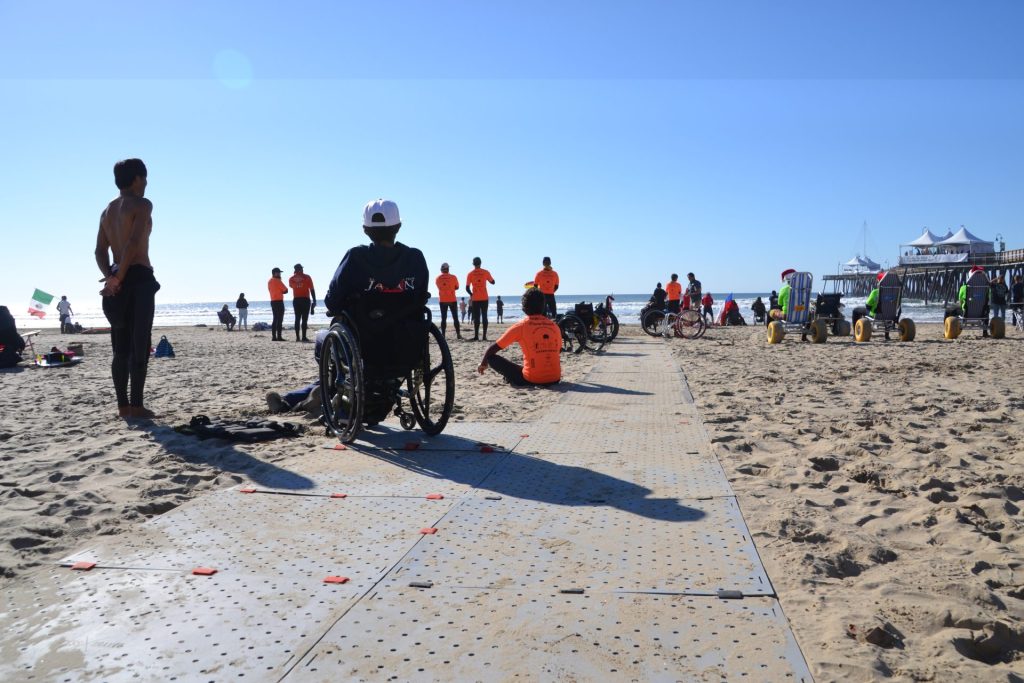 Bannière avec une plage avec du monde vu de dos et au milieu un tapis de plage large avec une personne en fauteuil roulant dessus