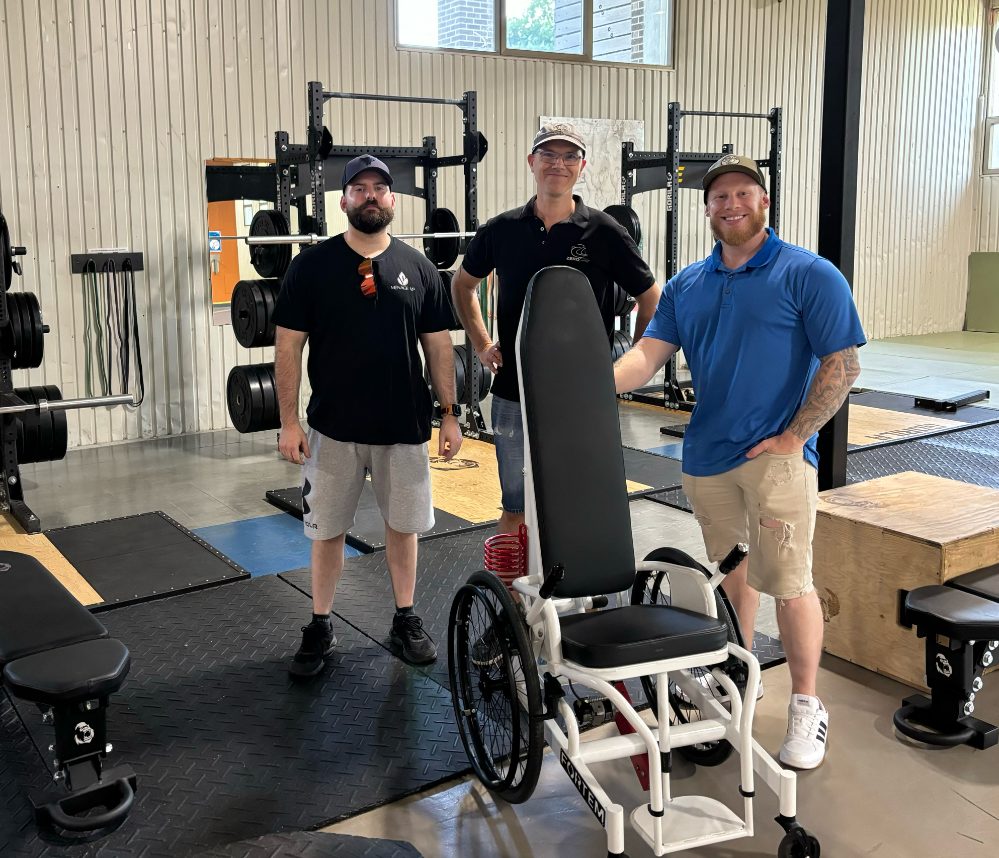 Trois personnes sont debout derrière un Gymchair, fauteuil roulant adapté pour l'entrainement