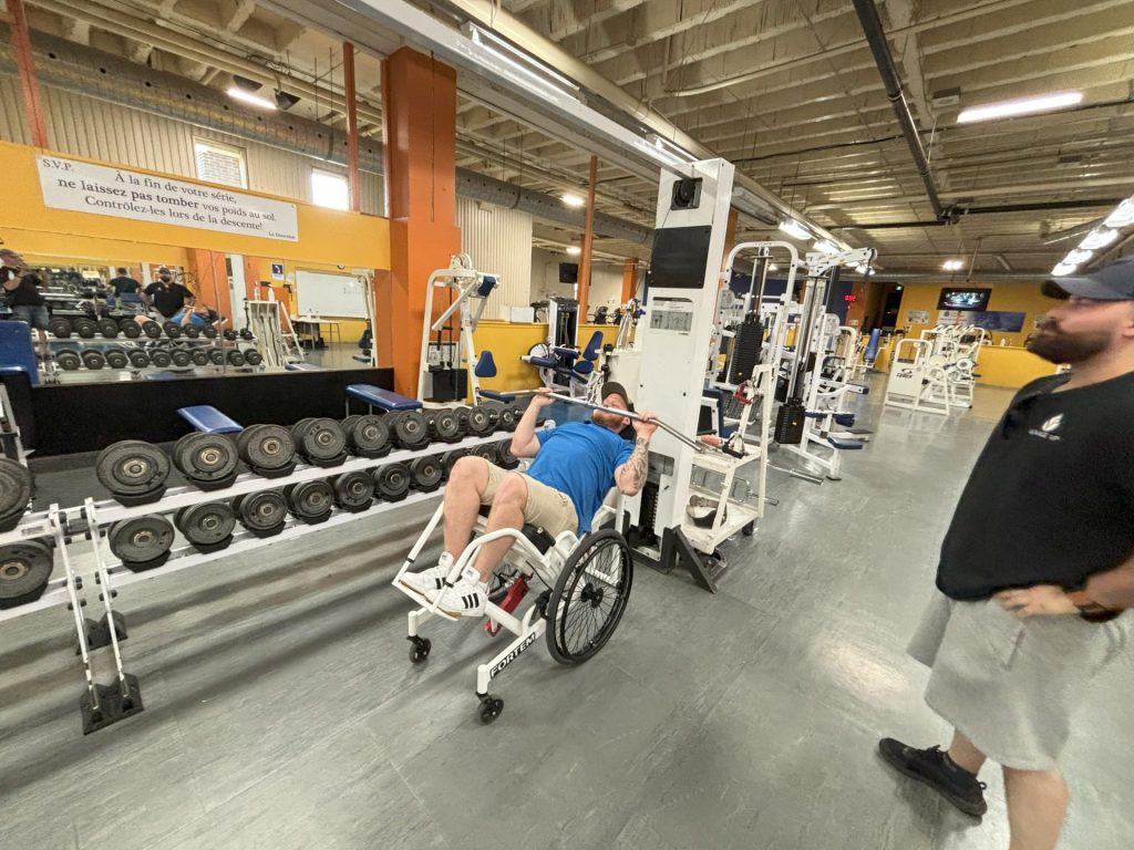 A man is on a wheelchair with a laid down backrest. He is in a gym, using a training machine. Un homme est en fauteuil roulant avec un dossier allongé. Il se trouve dans une salle de sport et utilise un appareil d'entraînement.