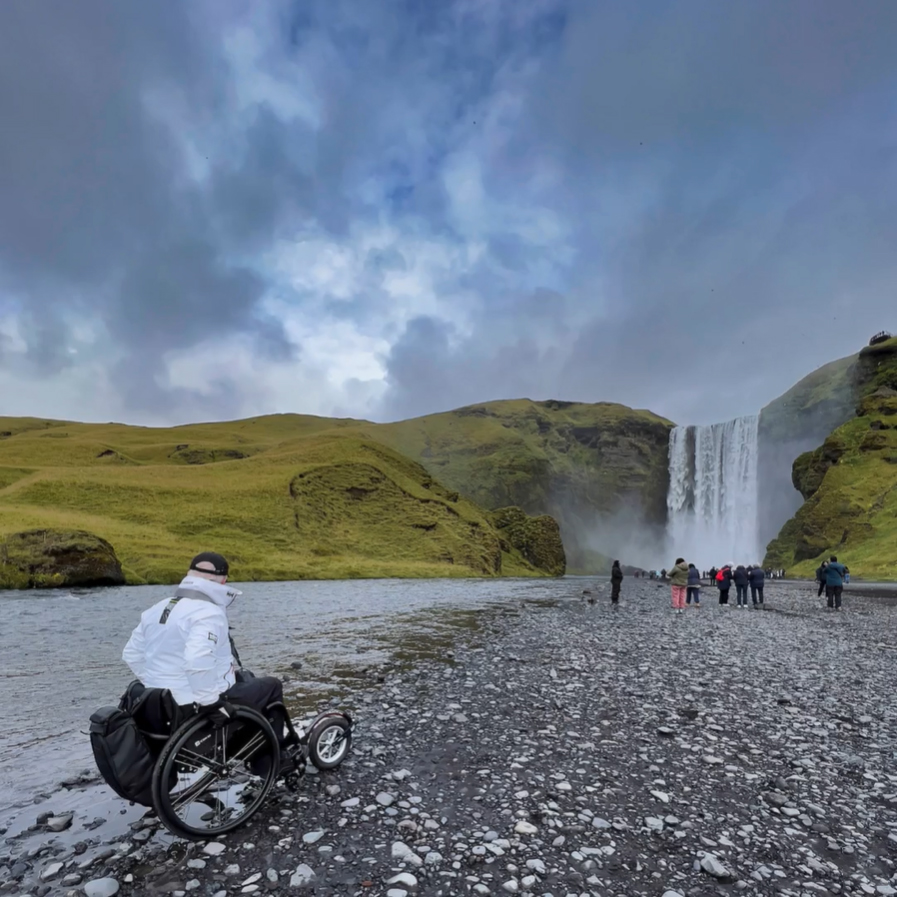 Une personne, vue de 3/4 arrière, est en fauteuil roulant avec un Track Wheel sur un chemin de roche, au bord d'une rivière et devant une grande cascade