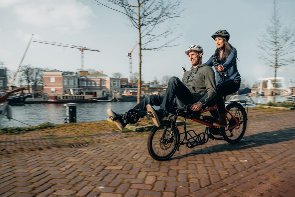 Deux personnes souriantes sont sur un tandem Pino, un homme à l'avant et une femme à l'arrière. Ils se trouvent dans une ville, sur une voie pavée au bord d'une rivière.