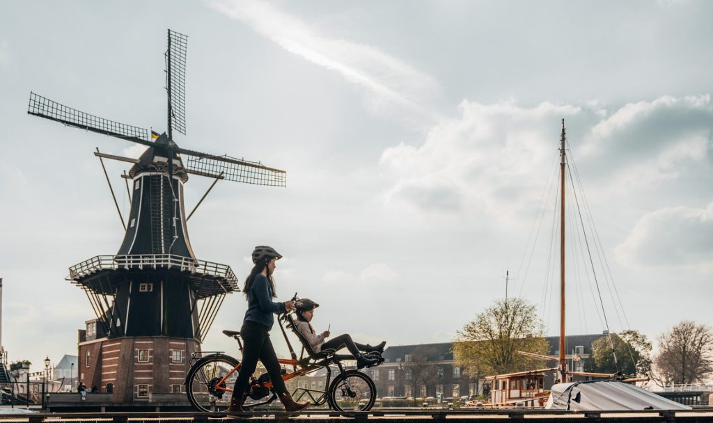 Deux personnes sont visibles de loin. Une jeune est assise devant un tandem Pino, l'autre, une femme, est debout à côté de la personne sur le vélo. Ils sont dans une ville, sur une jetée au bord de l'eau, avec un moulin à vent à l'arrière.