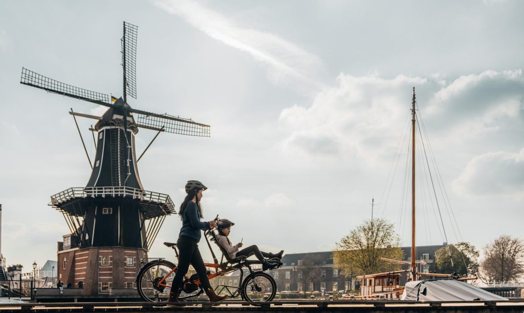 Two persons can be seen from afar. A young one is sitting in front of a Pino tandem bike, the other one, a woman, is standing next to the person on the bike. They are in a city, on a pier next to water and there is a windmill in the back.