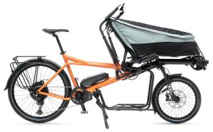 The photo shows an orange Pino tandem bike in cargo version, with a transport rack under the frame and a kind of large bag at the front. This is an electric version on a white background.