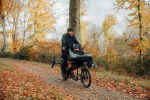 A man and child ride a Pino tandem on a country lane. The child is at the front, in a protective canvas. Both are smiling. There's another bike, the boy's bike, on the rear rack.