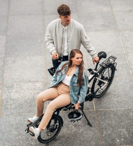 A young man and a young woman are with a Pino tandem bike in town, at a standstill. The young man is standing next to the bike and the young woman is sitting on the front seat, which is semi-recumbent.