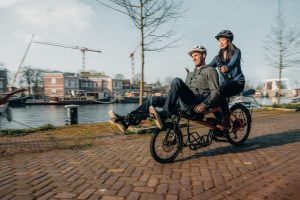 A woman and a man are on a Pino tandem bike with the woman in the back and the man in the front. They are on a paved street, by the water, in the city.