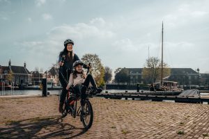 A young woman and a child ride in tandem on a Pino bike in town. They're all smiles as they ride along a paved riverside lane.