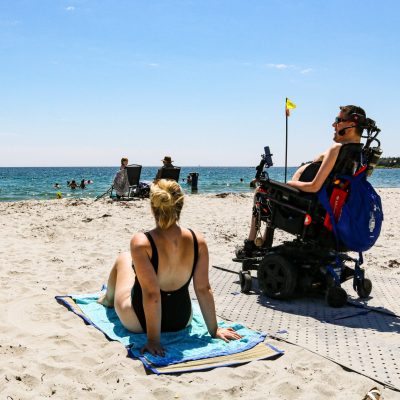 Deux personnes qui sont à la plage, l'une en fauteuil roulant électrique, sur des dalles de tapis de plage, et l'autre à côté, allongée sur une serviette