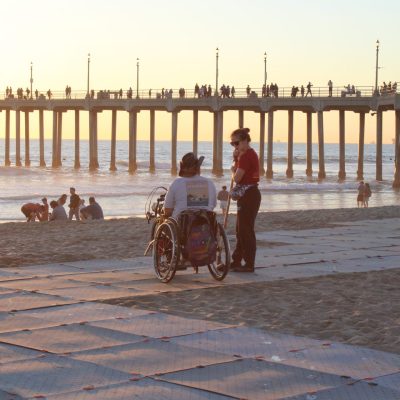 Deux personnes sur une plage devant un quai élevé au loin. Elles sont sur un Access Trax, une des personnes est en fauteuil roulant et l'autre est debout