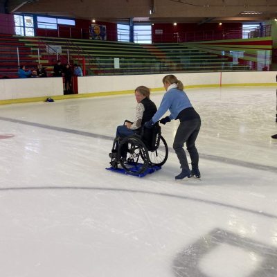 Deux femmes font du patin à glace dans un arena. L'une pousse la deuxième qui est dans un fauteuil roulant et sur un Lugicap Ice Perf / Two woman are ice-skating in an arena. The first one pushes the other one who is on a wheelchair which is on a Lugicap Ice Perf