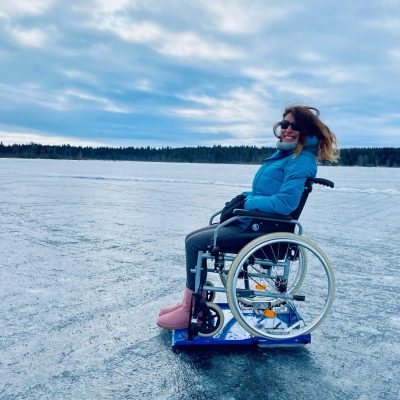 Une femme est sur un fauteuil roulant qui est sur un Lugicap Ice Perf sur un lac gelé / A woman is on a wheelchair which is on a Lugicap Ice Perf, on a frozen lake