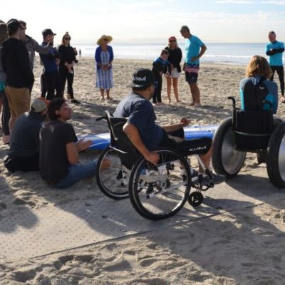 Groupe de personnes en fauteuil roulant et pas en fauteuil sur un tapis de plage, sur la plage